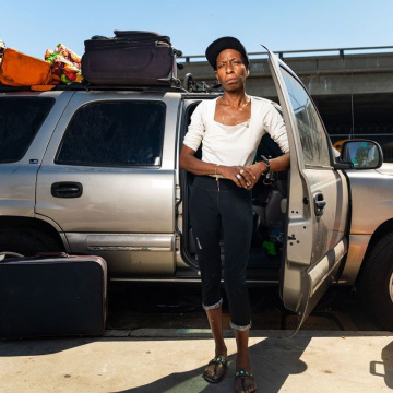 Portrait of a Black unhoused woman standing in front of her gray SUV, elbow propped against a door, her luggage stacked nearby
